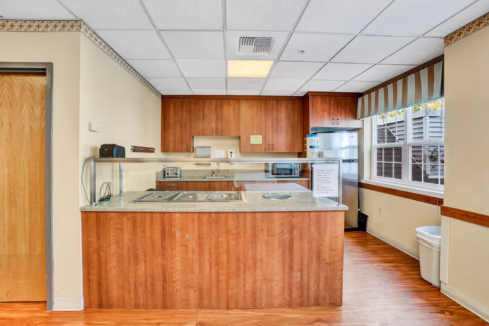 Kitchenette serving counter with wood cabinetry, stainless refrigerator, and a glass sneeze guard next to a window.