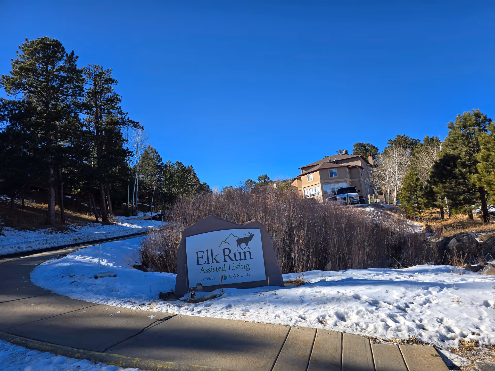 Outdoor view of Elk Run Assisted Living facility sign surrounded by snow and trees, with a building and clear blue sky in the background.