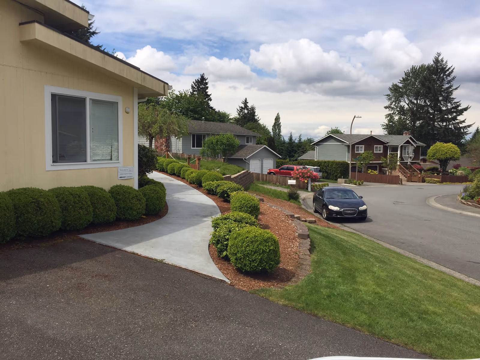 View of the exterior of a residential building with a paved walkway lined with neatly trimmed bushes leading to the entrance. The scene includes a street with parked cars and neighboring houses under a partly cloudy sky.