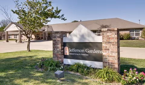 Exterior view of Jefferson Gardens Senior Living facility with a brick sign in the foreground displaying the facility name. The building and a tree are visible in the background under a clear sky.