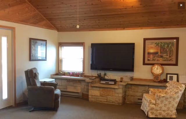 A cozy living room with a wooden vaulted ceiling, a large flat-screen TV mounted on a stone wall, a gray recliner chair on the left, a floral upholstered armchair on the right, a window with blinds and a small table beneath it, framed artwork on the walls, and a wooden clock on a shelf.