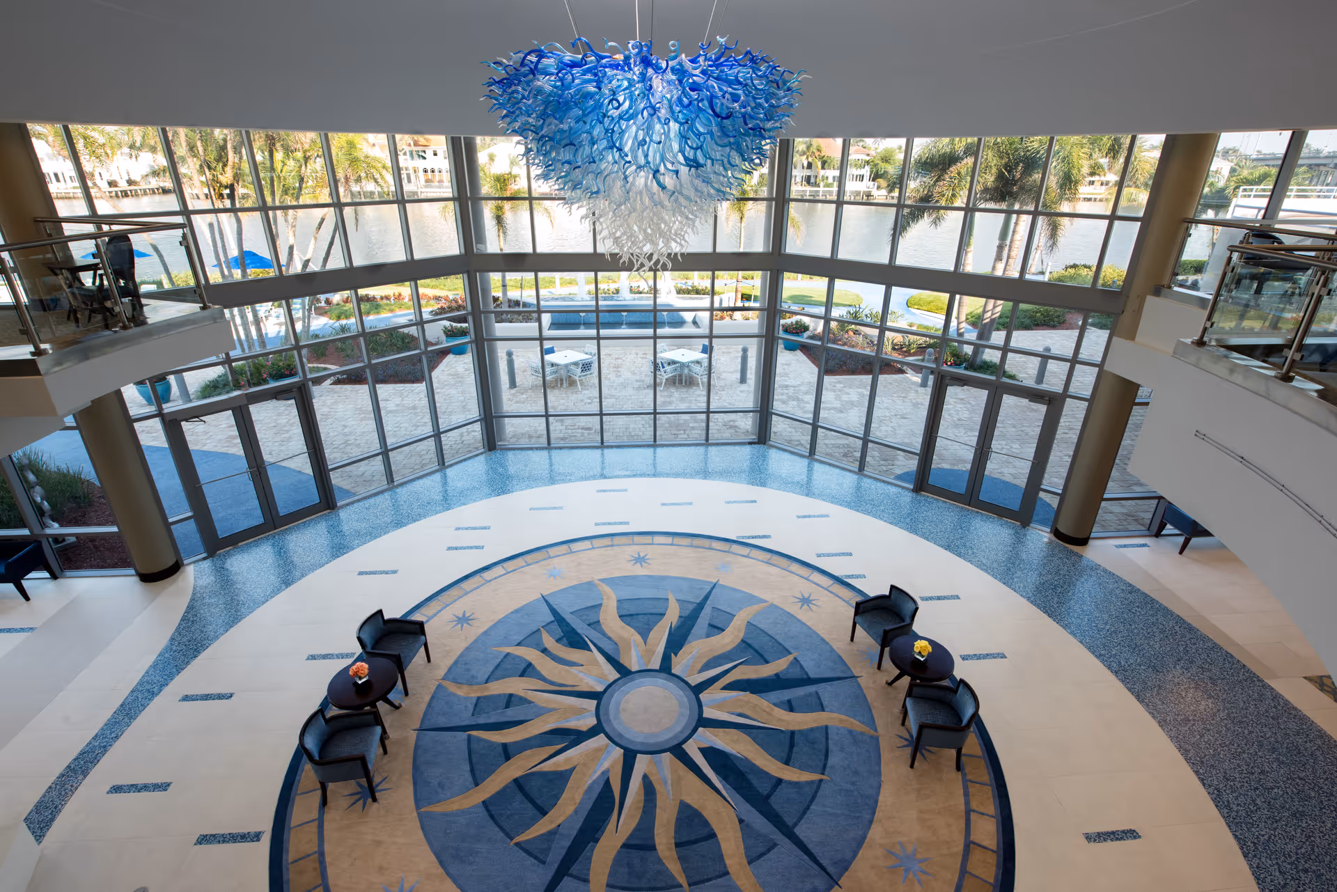 Spacious two-story lobby with a large circular floor medallion, seating, a blue glass chandelier, and floor-to-ceiling windows overlooking water.