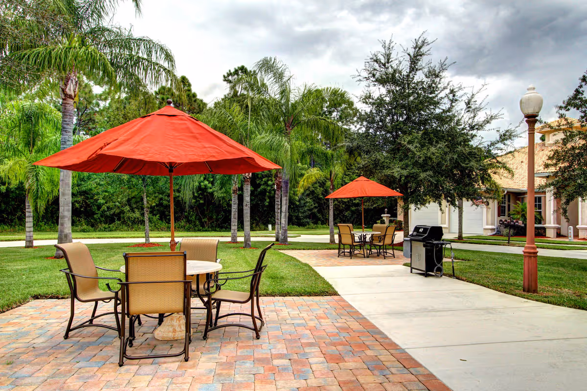 Outdoor patio area with two sets of tables and chairs under red umbrellas, surrounded by palm trees and greenery. A barbecue grill is placed near the sidewalk, and a building with a porch is visible in the background under a cloudy sky.