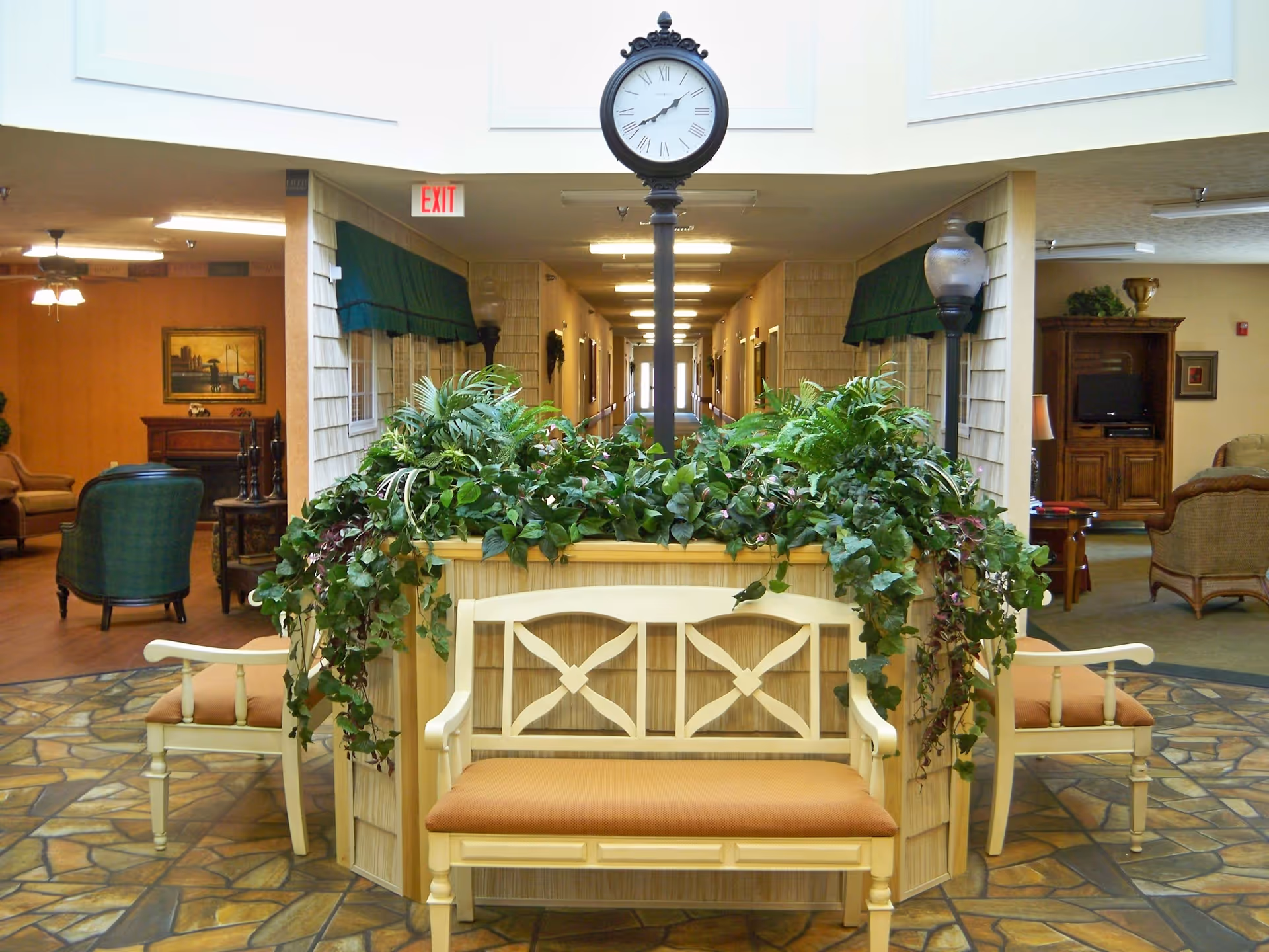 Interior view of a retirement community common area featuring a central planter with green leafy plants and a vintage-style clock on a pole. Surrounding the planter are cushioned wooden benches. In the background, there is a hallway with doors on either side and seating areas with chairs and a TV cabinet on both sides of the image.