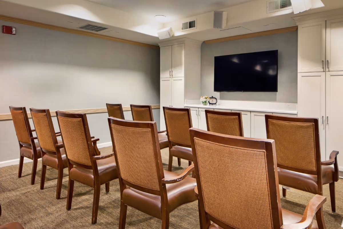 Small screening room with rows of wooden chairs facing a wall-mounted flat-screen TV and built-in cabinets.