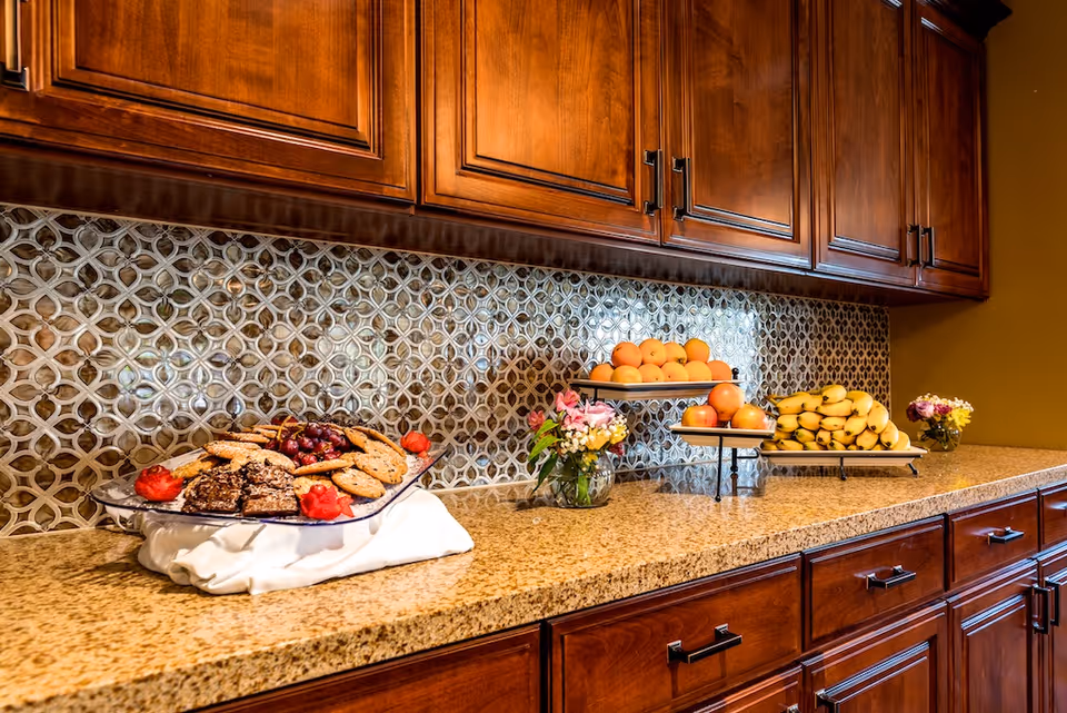 Granite countertop buffet with plates of fruit and pastries, small flower arrangements, wooden cabinets, and a decorative tile backsplash.