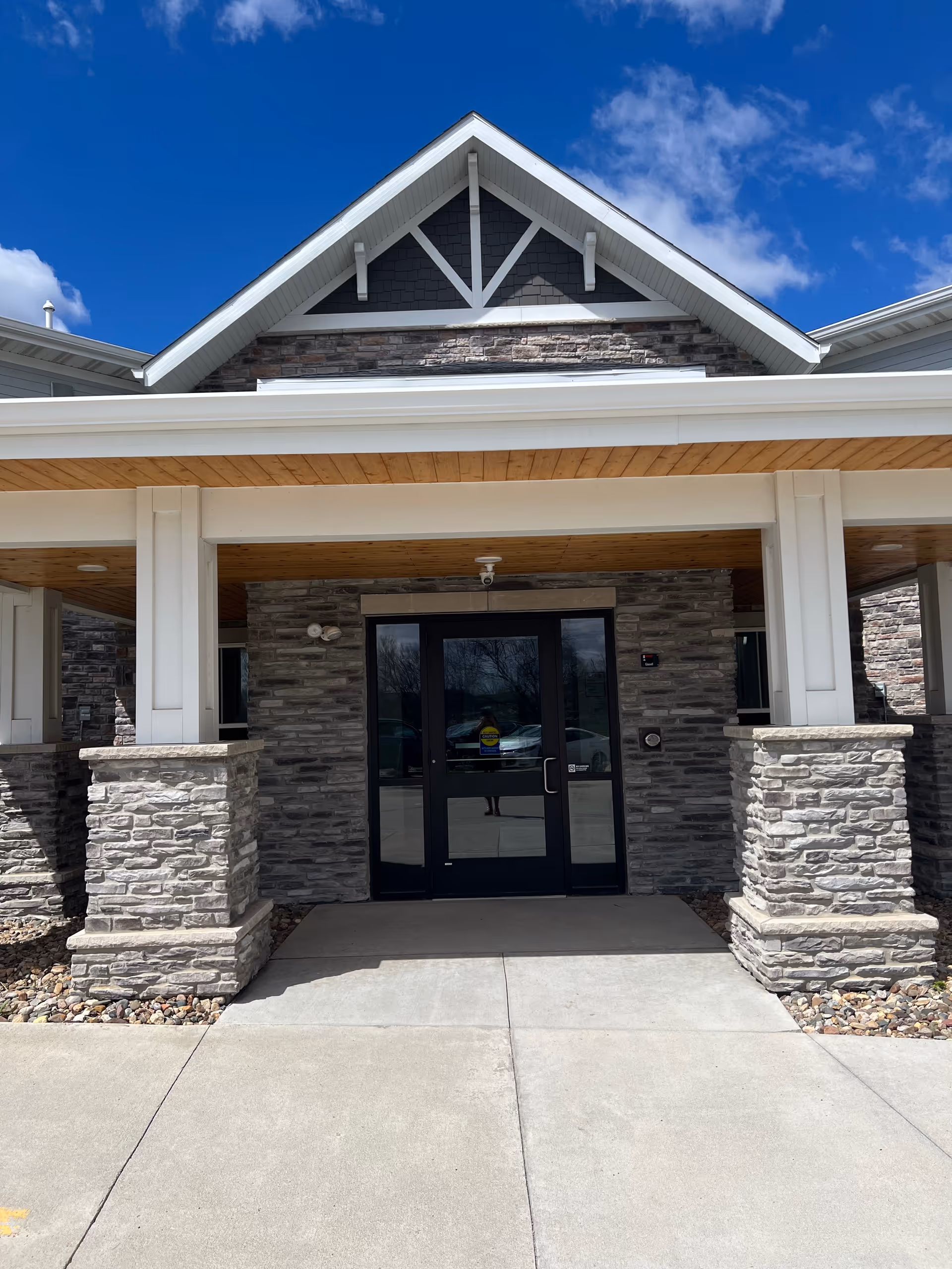 Covered stone and wood building entrance with double glass doors under a blue sky.