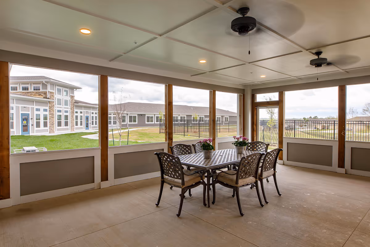 Covered screened patio with a metal dining table, six chairs, ceiling fans, and views of the facility courtyard.