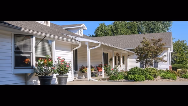 Exterior view of a single-story building with white siding and a gray roof, featuring a covered entrance with white pillars, potted plants, and shrubs along the walkway under a clear blue sky.