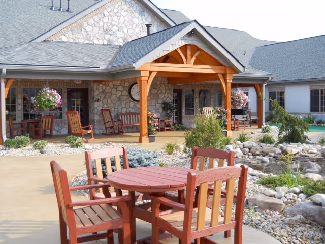 Outdoor patio area with a red wooden table and chairs in front of a covered porch and landscaped rock garden at a senior living facility.