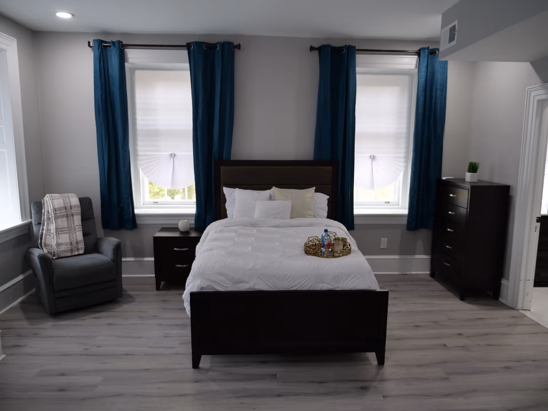 A neatly arranged bedroom with a dark wooden bed frame and white bedding, positioned between two windows with blue curtains and white blinds. To the left of the bed is a gray armchair with a plaid blanket draped over it, and a small dark wooden nightstand with a decorative item. To the right is a dark wooden chest of drawers with a small green plant on top. The room has light gray walls and light wood flooring.