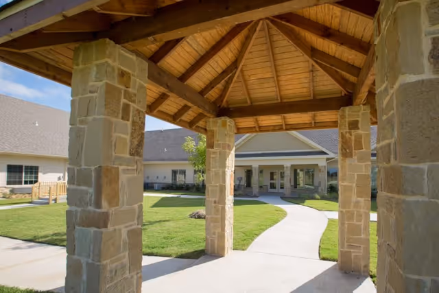 View from under a wooden gazebo with stone pillars looking out onto a paved walkway leading to the entrance of a single-story building with a pitched roof, surrounded by green grass and landscaping.