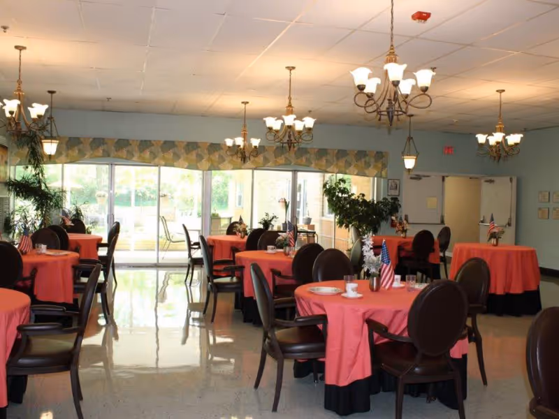 A dining room with multiple round tables covered with red tablecloths and black chairs around them. Each table has small decorations including American flags and flowers. The room has large windows and glass doors letting in natural light, with several chandeliers hanging from the ceiling. There are plants near the windows and a door at the back of the room.