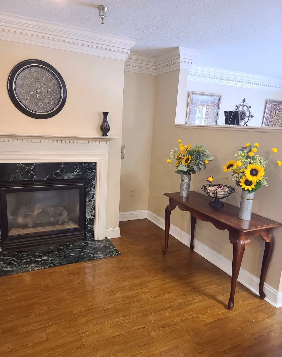 Cozy living room corner with a marble fireplace, wall clock, and wooden console table topped with sunflower arrangements.
