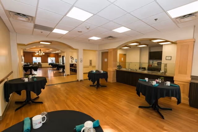 Dining room with round tables set with cups and napkins, wood floors, archways, and a serving counter in the background.