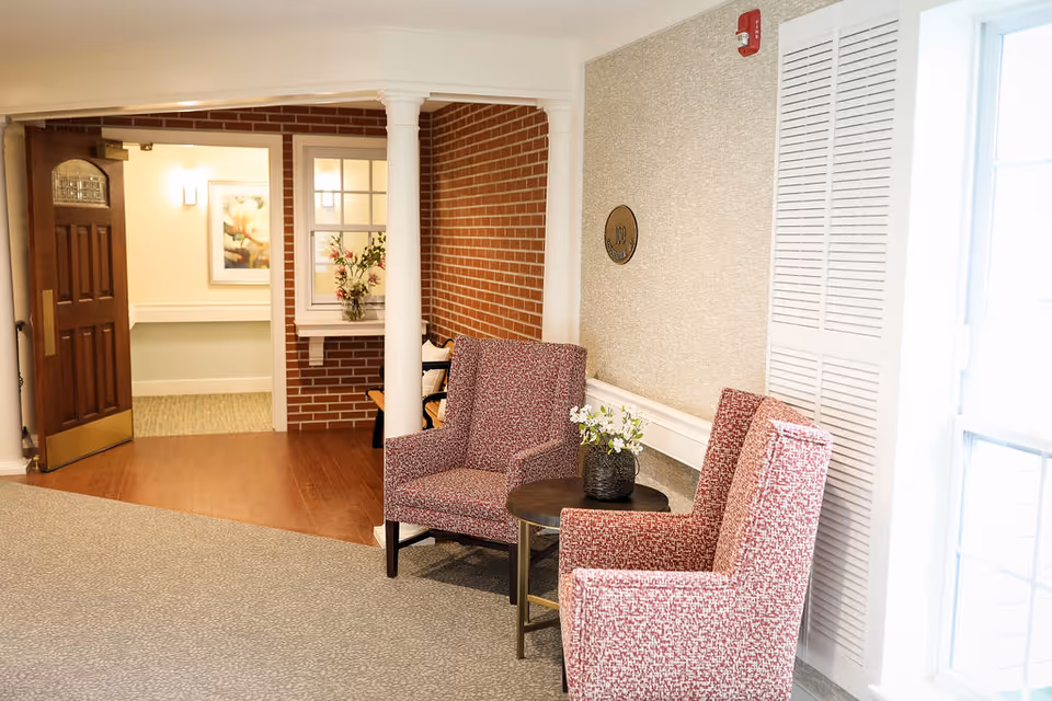A cozy seating area in a senior living facility featuring two patterned armchairs and a small round table with a flower vase. The space has a mix of carpet and wood flooring, a brick accent wall, white columns, and a wooden door leading to another room with framed artwork and soft lighting.