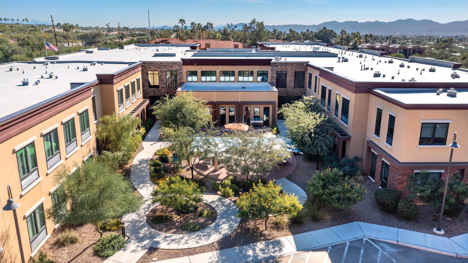 Aerial view of The Watermark at Oro Valley senior living facility showing a U-shaped building surrounding a landscaped courtyard with walking paths, trees, and shrubs. The building has two stories with multiple windows and a flat roof. Mountains and desert vegetation are visible in the background under a clear blue sky.