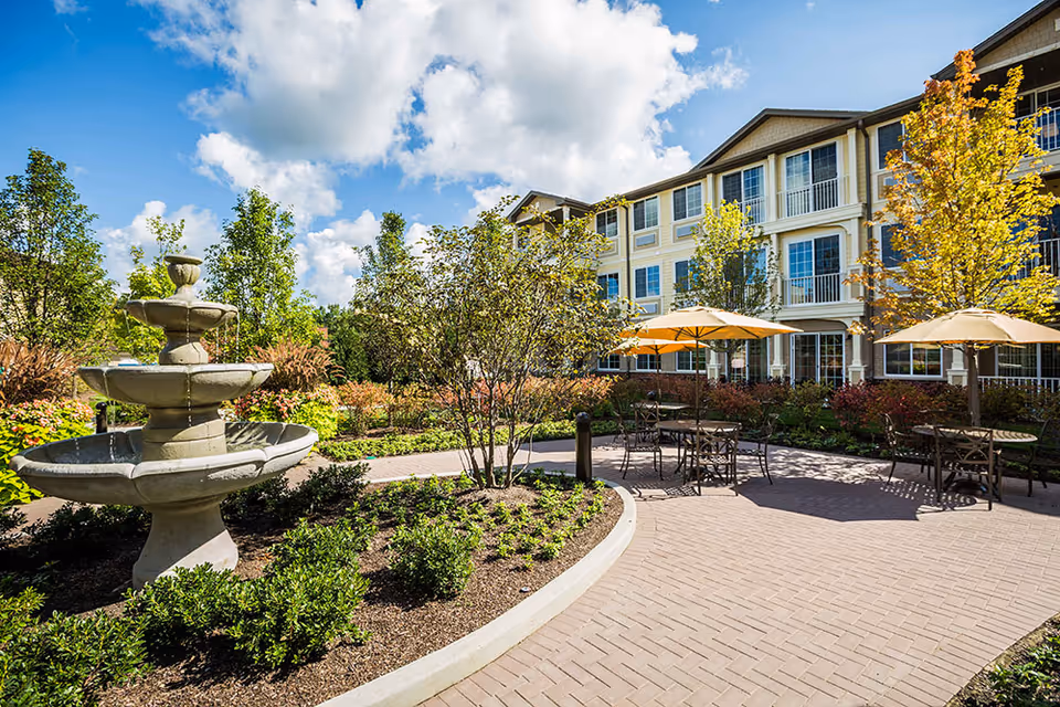 Outdoor patio area at Westbrook Senior Living featuring a multi-tiered stone fountain, landscaped garden beds with shrubs and small trees, several tables with chairs under yellow umbrellas, and a multi-story building in the background under a partly cloudy blue sky.