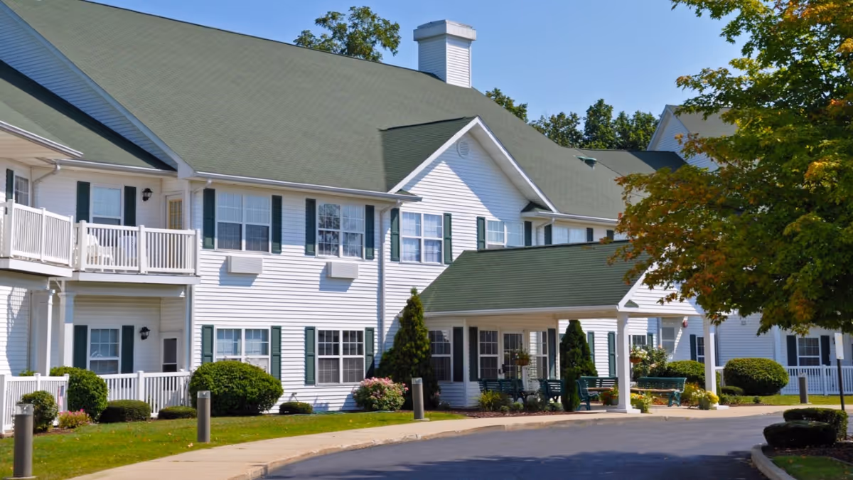 Exterior view of Oak Woods Manor, a two-story white building with green shutters and a green roof. The entrance features a covered porch with seating, surrounded by well-maintained landscaping including bushes, flowers, and a tree with green and orange leaves.