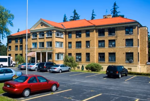 Three-story brick building with a red roof and a parking lot with cars in front.