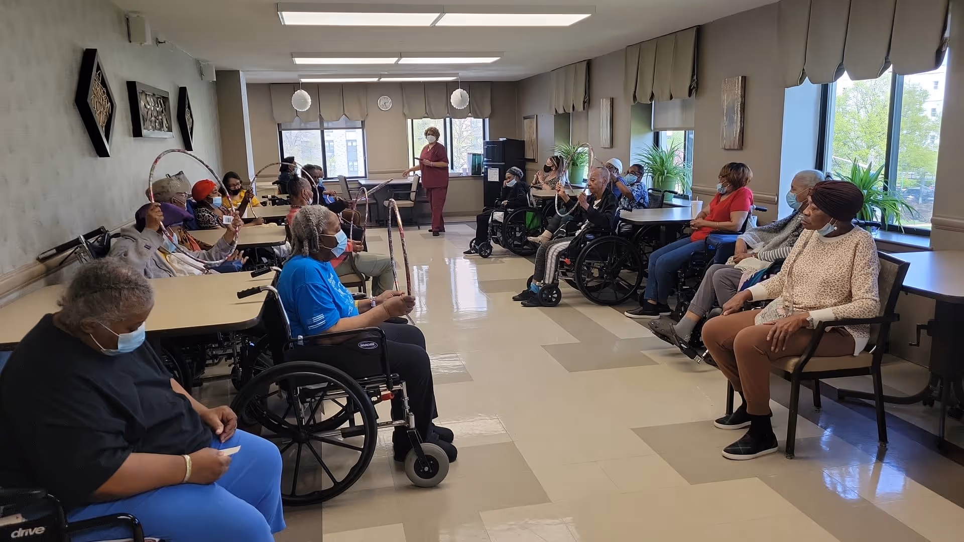 A group of elderly individuals, some in wheelchairs and others seated in chairs, participate in a seated exercise or activity session in a bright room with large windows. A staff member stands at the front of the room leading the activity. The participants are spaced along the walls, holding exercise hoops or similar equipment, and many are wearing face masks.