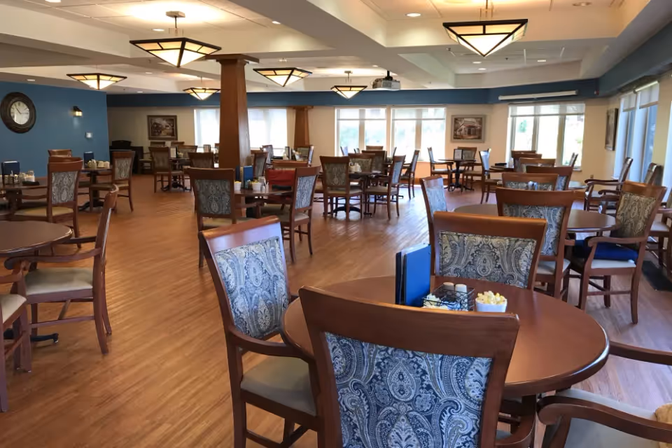 Spacious dining room with multiple round wooden tables and patterned chairs, hardwood floors, pendant lights, and large windows.