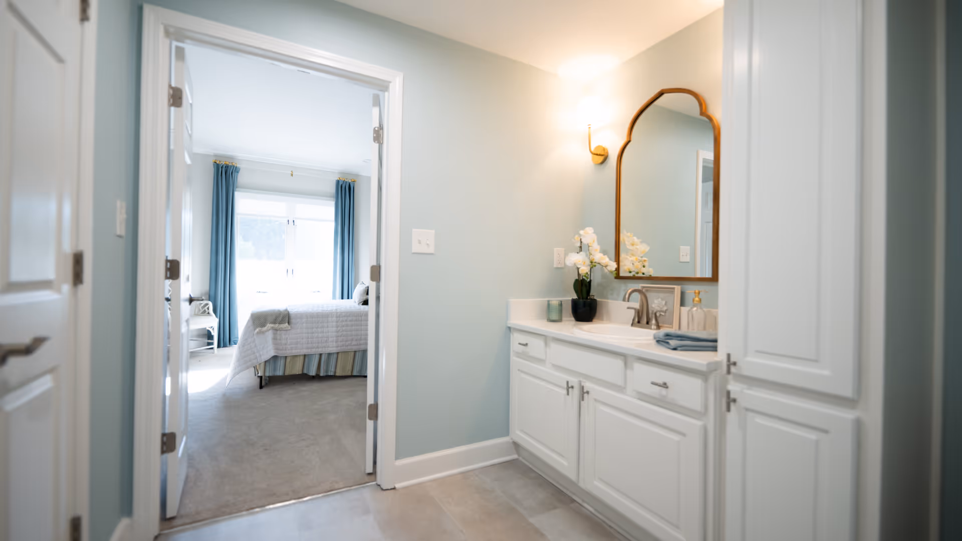 View of a bathroom vanity area with a large mirror, white cabinets, and a sink. A wall sconce light is mounted above the mirror. Through an open doorway, a bedroom with a bed, blue curtains, and a window letting in natural light is visible.