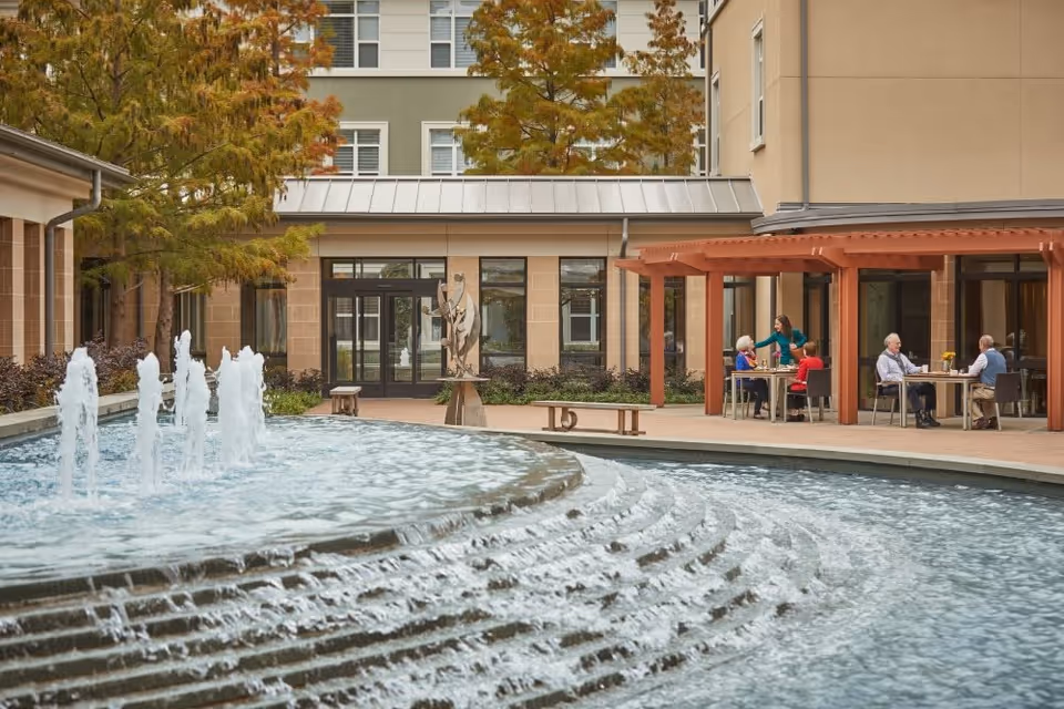 Outdoor courtyard area at The Legacy Willow Bend featuring a large tiered water fountain with multiple water jets. In the background, there are trees with autumn foliage, a building with large windows, and a covered patio where several elderly people are seated at tables, engaging in conversation and being served by a caregiver.