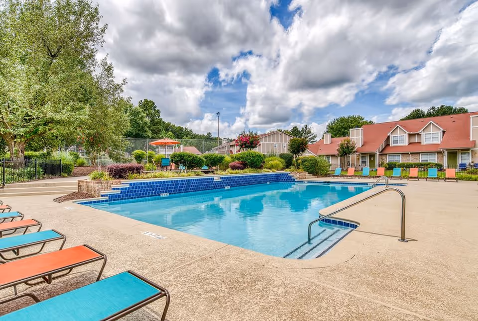 Outdoor swimming pool with blue water surrounded by a concrete deck with colorful lounge chairs. In the background, there are trees, shrubs, and residential buildings with red roofs under a partly cloudy sky.
