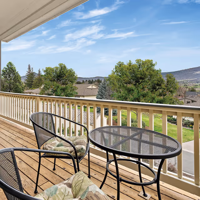 A balcony with a wooden floor and beige railing, featuring two black metal chairs with floral cushions and a matching round metal table. The balcony overlooks a scenic view of trees, rooftops, and distant hills under a partly cloudy blue sky.