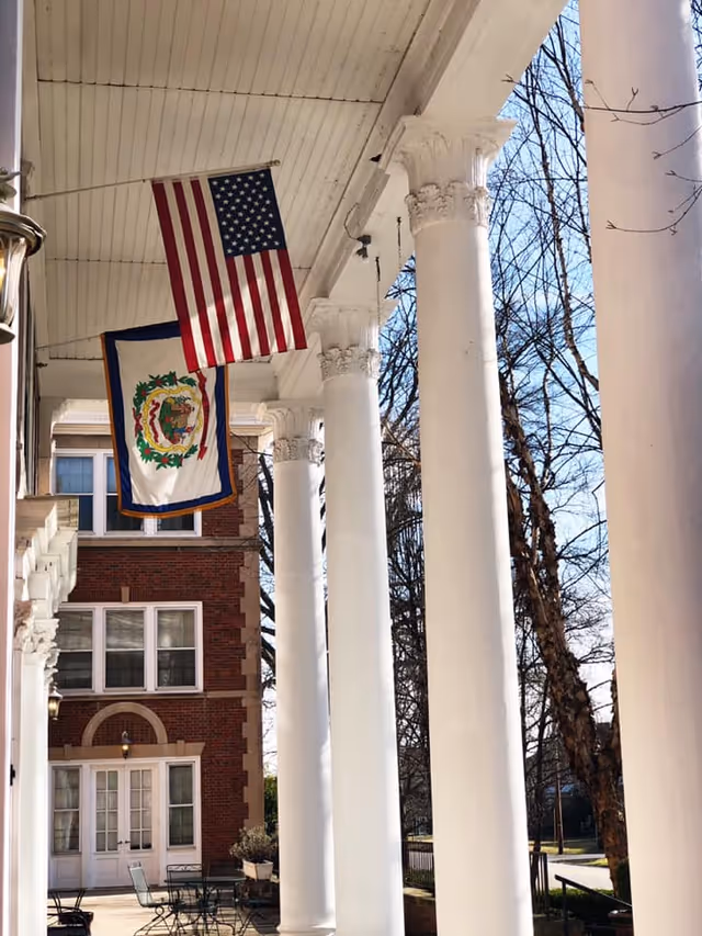 View of a building's front porch with tall white columns and two flags hanging from the ceiling, one American flag and one West Virginia state flag. The building is made of red brick with white trim around the windows and doors. Leafless trees and a clear sky are visible in the background.