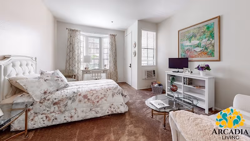 A bright and cozy bedroom with a floral-patterned bedspread on a white tufted headboard bed, a glass side table, a white desk with a chair near a window with patterned curtains, a white TV stand with a small TV and decorative items, a glass coffee table, and a comfortable chair with a fluffy throw. A colorful painting hangs on the wall, and there is a window air conditioning unit. The room has brown carpet flooring and white walls.