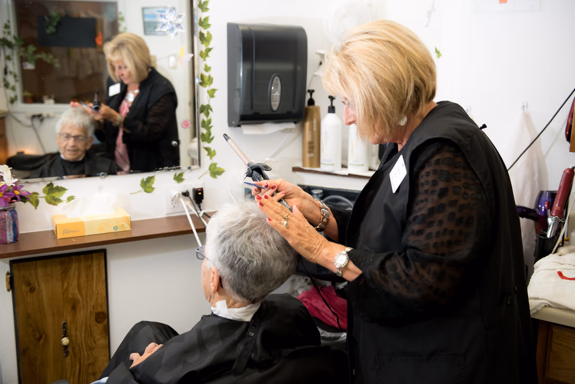 A hairstylist is curling the hair of an elderly woman seated in a salon chair. The scene is reflected in a mirror decorated with green leaf garlands and paper snowflakes. Various hair care products and a tissue box are visible on the counter.