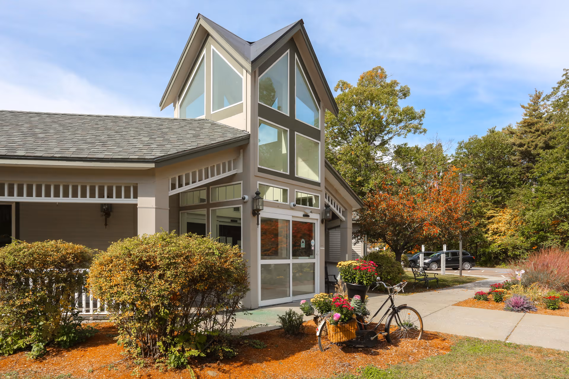 Exterior view of a senior living facility building with large windows and a peaked roof. There are bushes and a decorative bicycle planter with flowers near the entrance. Trees with autumn foliage and parked cars are visible in the background under a partly cloudy sky.