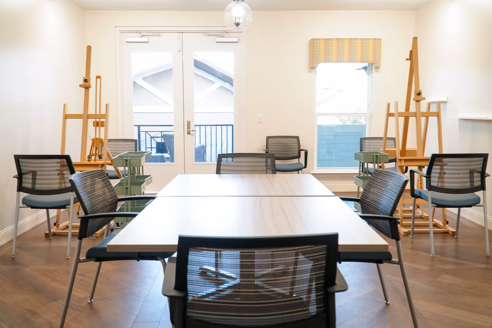A bright room with a rectangular table surrounded by six black mesh chairs. There are wooden easels and green rolling carts positioned around the room. A glass door and a window with a colorful valance allow natural light to enter the space.