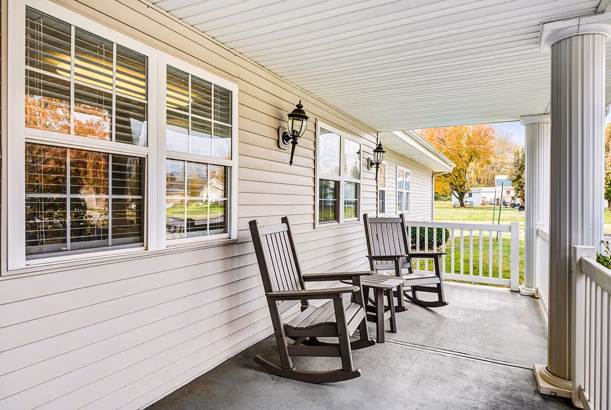 Covered porch area with two wooden rocking chairs and a small table between them, beige siding on the building, white columns, and a view of green grass and trees with autumn foliage in the background.