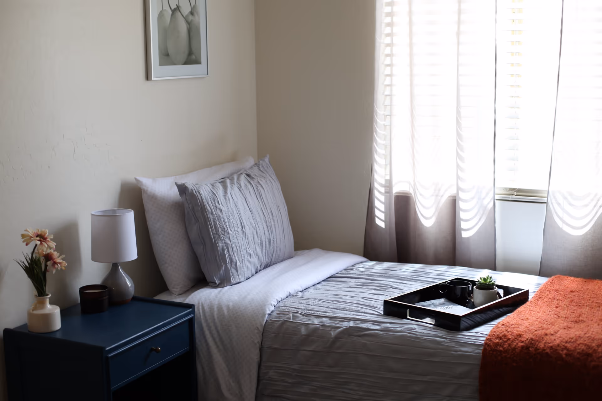 A neatly made single bed with gray and white bedding next to a window with sheer curtains. A black tray with a cup and a small plant sits on the bed. Beside the bed is a dark blue nightstand with a white lamp, a small candle, and a vase with flowers. A framed picture of pears hangs on the wall above the bed.