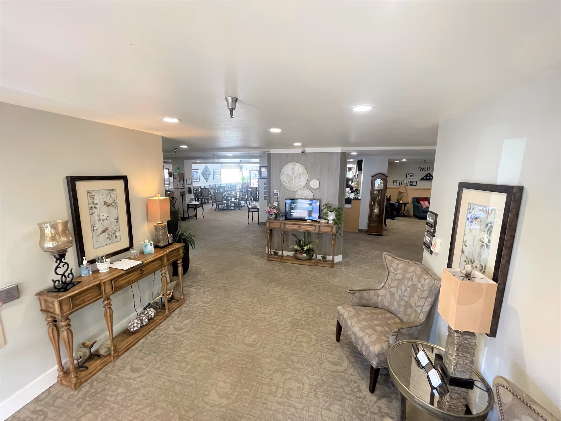 Interior view of a senior living facility hallway with beige carpet and walls. There are decorative tables with lamps, framed artwork, and plants along the walls. In the background, there is a dining area with multiple tables and chairs, and a living room area with a grandfather clock and seating.