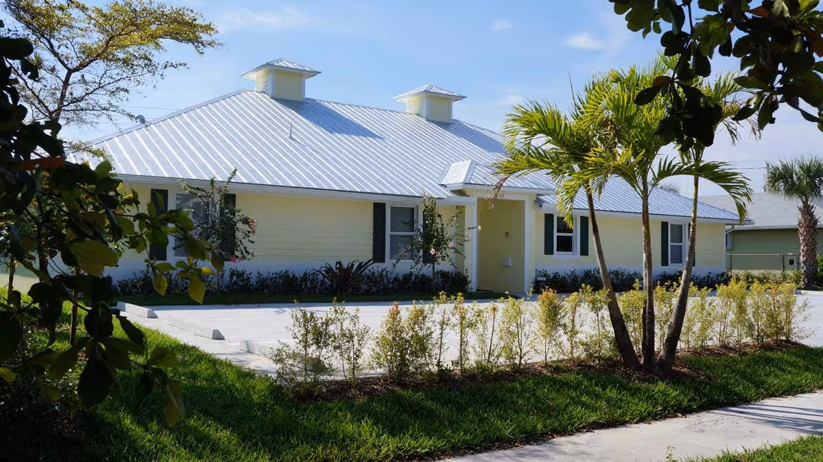 Single-story light-yellow building with a metal roof, palm trees, landscaping, and a paved parking area in front.