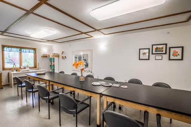 A well-lit room with a long black table surrounded by black chairs. On the table, there is a vase with orange and white flowers and several small plates or coasters. The room has white walls with framed artwork and a large window with a green valance letting in natural light. The ceiling has wooden trim and rectangular light fixtures.