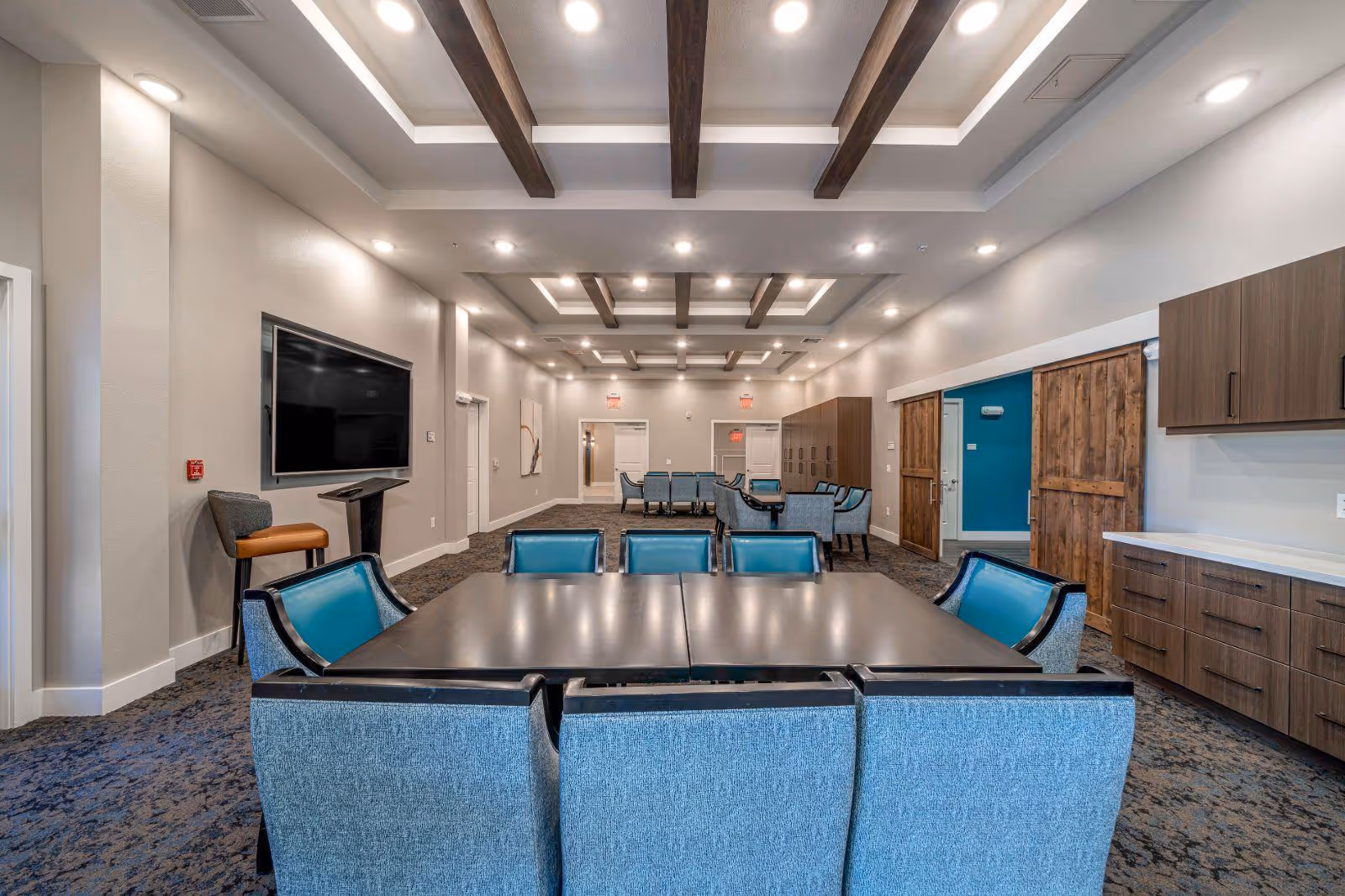 Spacious communal dining/activity room with dark tables, teal-upholstered chairs, a wall-mounted TV, recessed beamed ceiling, and built-in cabinetry.