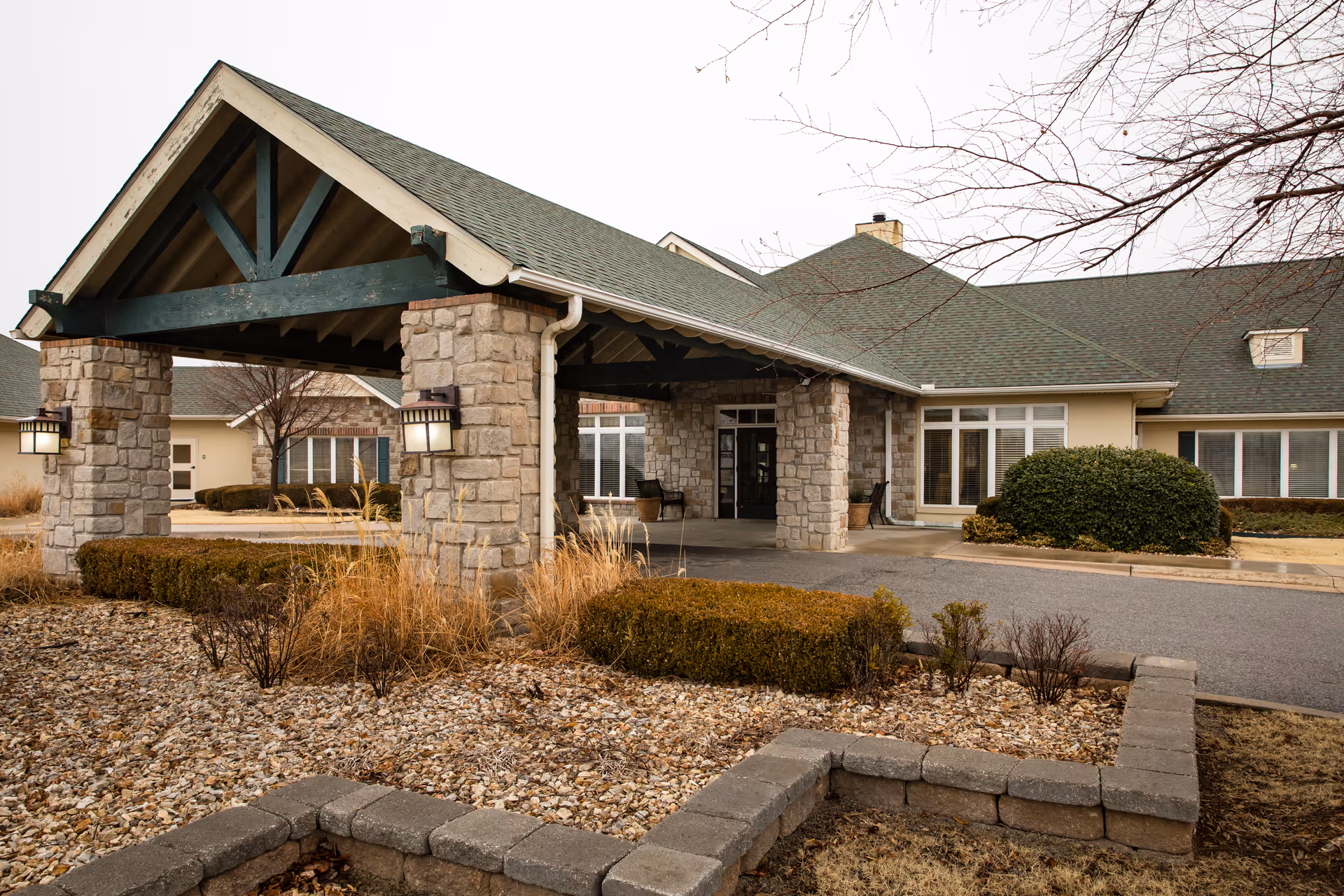 Exterior view of a senior living facility entrance with a covered driveway supported by stone pillars, landscaped with bushes and dry grass, and a building with large windows and a green roof in the background.