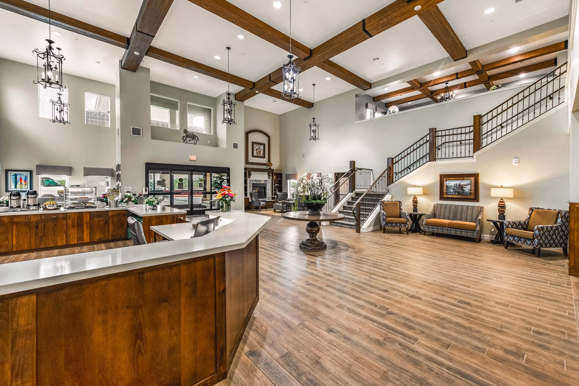 Bright, open senior-living lobby with a wooden reception desk, seating areas, a central table with flowers, and a staircase under exposed wooden ceiling beams.
