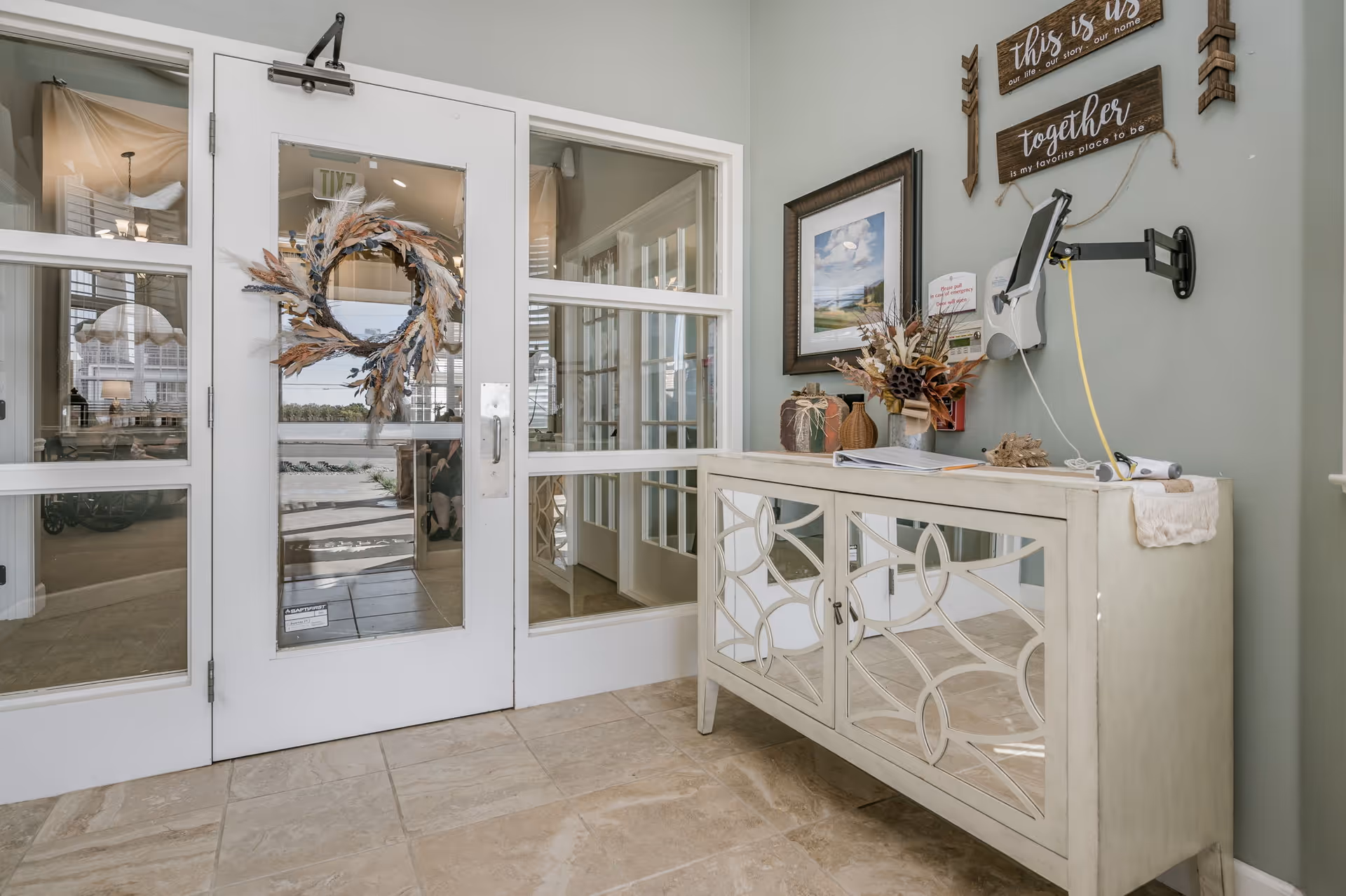 Interior view of a hallway or entrance area in a senior living facility with a white door decorated with a wreath made of dried flowers and leaves. To the right, there is a mirrored cabinet with decorative items including vases and dried floral arrangements. Above the cabinet, there are two wooden signs with inspirational text and a mounted tablet device on an adjustable arm. The floor is tiled and the walls are painted light gray.