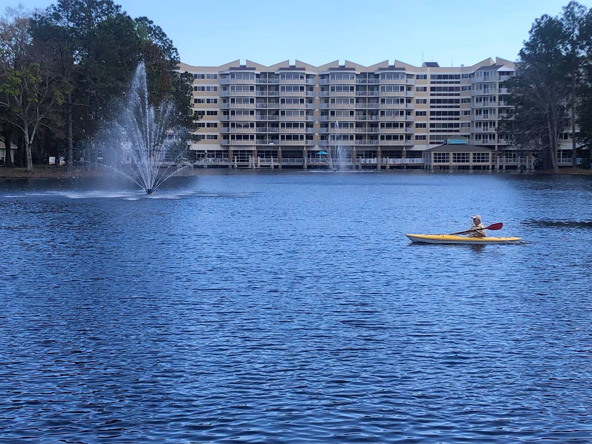A large multi-story building with balconies overlooks a lake with two water fountains. A person is kayaking on the lake in a yellow kayak. Trees surround the lake on both sides.