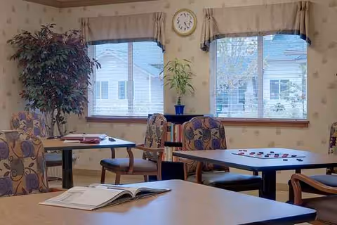 Sunlit dining room with tables, chairs, plants, a clock, and magazines.