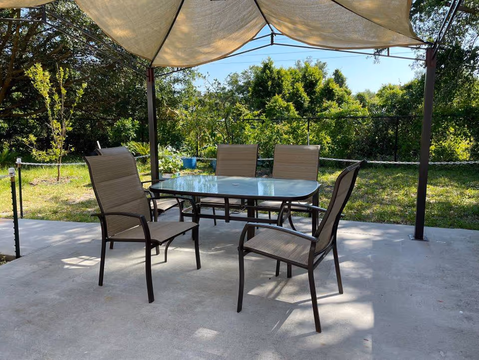 Outdoor patio with a glass-top dining table and several chairs under a fabric canopy, with trees and grass in the background.