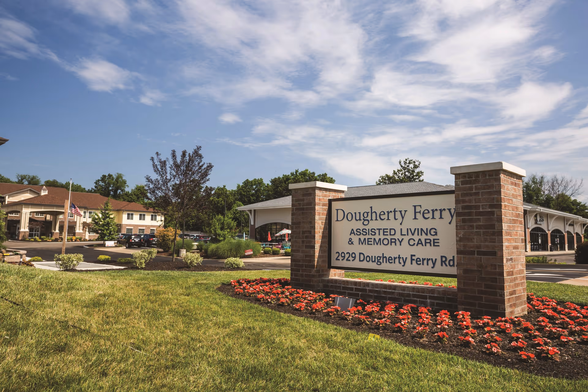 Outdoor view of Dougherty Ferry Assisted Living and Memory Care facility with a large sign displaying the facility name and address, surrounded by a flower bed and green lawn under a partly cloudy sky.