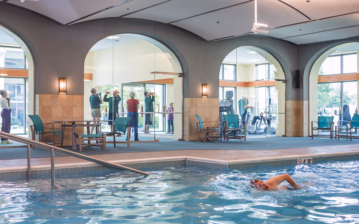 Indoor swimming pool area with a person swimming in the pool. In the background, there are large arched windows and glass walls showing a fitness room where several elderly people are exercising and drinking water. The area has chairs and tables along the poolside.