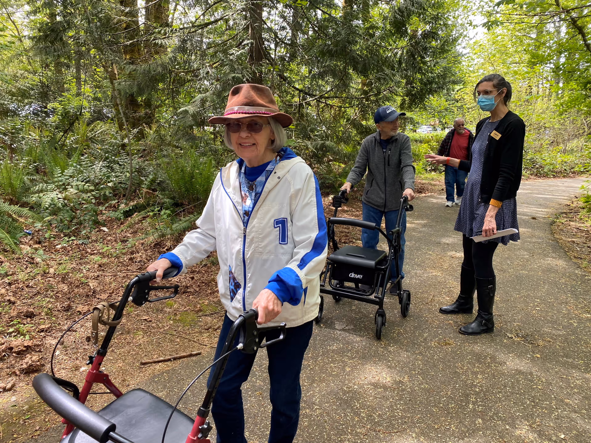 An elderly woman wearing a brown hat and white jacket is walking with a red walker on a paved path surrounded by greenery. Behind her, an elderly man with a blue cap and gray jacket uses a black walker. A woman wearing a face mask, black cardigan, and dress is talking to them while holding a notebook. Another elderly man walks further behind on the path.